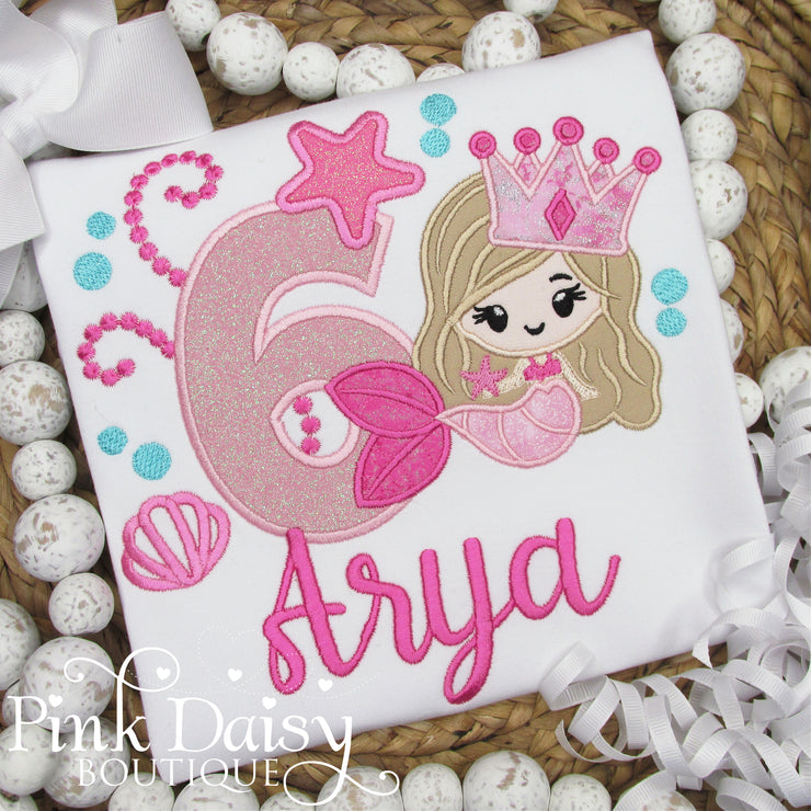 A personalized white birthday shirt for a young girl, featuring an embroidered pink mermaid with flowing hair, a glittery crown, and a smiling face. The mermaid sits beside a large pink number 6, accented with sparkly details. Below the design, the name "Arya" is stitched in hot pink thread. The shirt is displayed on a woven background and surrounded by white speckled beads and white ribbon curls, creating a festive and whimsical presentation.