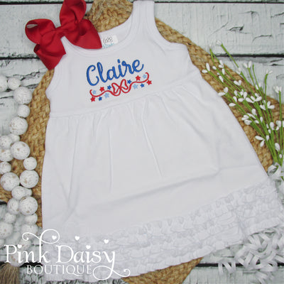 White sleeveless toddler girl dress with embroidered name Claire in blue script, red stars, and blue swirls, featuring a ruffled hem. Perfect for 4th of July celebrations, summer birthdays, patriotic parties, and toddler photo shoots. Handmade with love and personalized for a special touch.